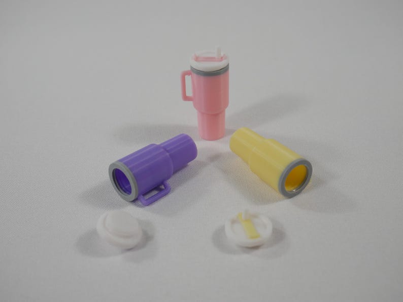 May include: A collection of colorful, miniature plastic drinkware, including a pink tumbler with a handle and straw, two small purple and yellow tubes, and two white lids. The items are arranged on a white surface.