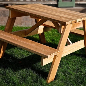 May include: A wooden picnic table with two benches. The table and benches are made of natural wood and have a rustic finish. The table is set on a grassy lawn.