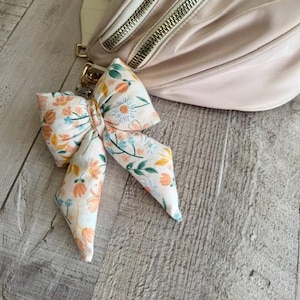 May include: A light pink fanny pack with gold zippers and a matching strap. A floral bow keychain with orange, blue, and white flowers hangs from the zipper. The background is a light wood surface.