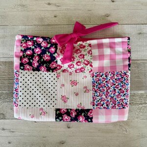May include: A patchwork fabric item with a pink bow. The item features a variety of floral and polka dot patterns in pink, white, and navy blue. The edges are trimmed with a pink gingham pattern.
