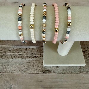 May include: Five beaded bracelets displayed on a stand. The bracelets feature a variety of small round beads in shades of cream, soft pink, metallic gold, and dark gray. Each bracelet incorporates different patterns of these colored beads.