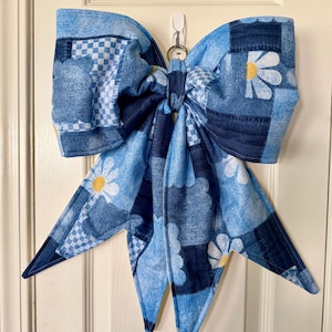 May include: A large fabric bow featuring a patchwork design in various shades of blue. The pattern includes white checkered squares, denim-like textures, and white daisy flowers with yellow centers. The bow has two long, pointed tails hanging down.