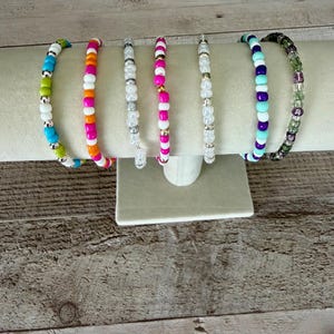 May include: A set of seven beaded bracelets displayed on a stand. The bracelets feature various color combinations including bright pink, orange, white, lime green, turquoise, purple, and translucent green beads, accented with small metallic silver beads.
