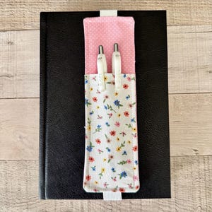 May include: A fabric pen holder with an elastic band attached to a black notebook. The holder features a pink and white polka dot upper section and a white lower section with a colorful floral pattern. Two white pens are inserted into the holder.