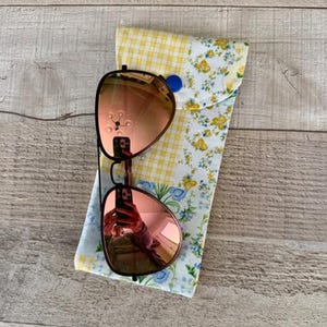May include: Rose-tinted sunglasses with a black frame rest on a floral and gingham patterned glasses case. The case is a mix of yellow gingham and a floral print with blue and yellow flowers. A blue button secures the case.