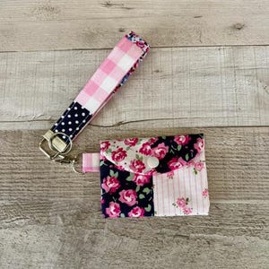 May include: A handmade fabric wristlet and matching small wallet. The wristlet features a pink and white gingham pattern, a navy blue polka dot pattern, and a floral pattern. The wallet has a floral pattern with a white button closure.