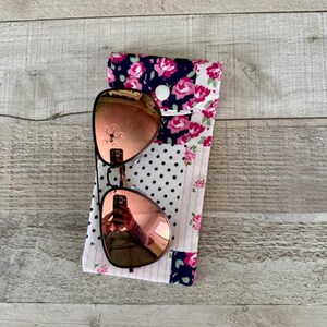 May include: Rose-tinted sunglasses rest on a floral print glasses case. The case features a patchwork design with pink roses on a navy background, white polka dots, and a striped pattern. A white button secures the case.