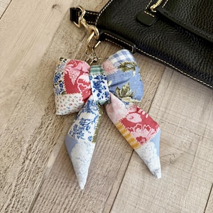 May include: A fabric bow bag charm featuring a patchwork design with various floral, gingham, and toile patterns in shades of red, blue, white, and green. The bow is attached to a gold-toned metal lobster clasp and ring, hanging from a black leather bag.