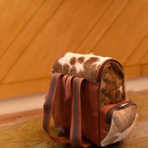 May include: A brown leather backpack with a cowhide pattern. The backpack has two adjustable straps and a zipper closure.
