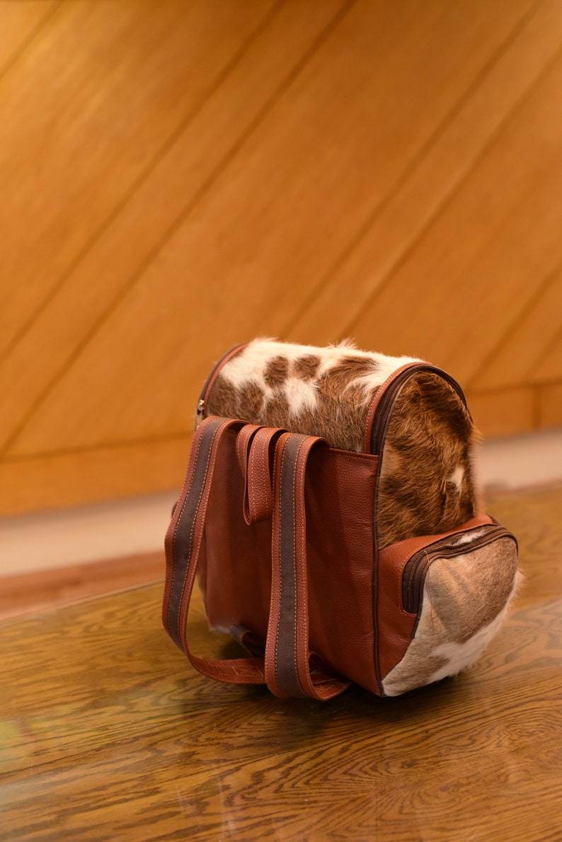 May include: A brown leather backpack with a cowhide pattern. The backpack has two adjustable straps and a zipper closure.