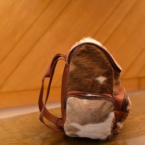May include: A brown leather backpack with a fur-covered front panel. The fur is a mix of brown and white.