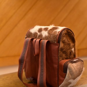 May include: A brown leather backpack with a cowhide flap and two brown straps. The backpack has a small pocket on the front.