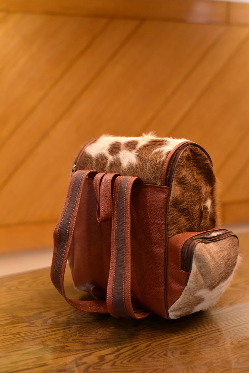 May include: A brown leather backpack with a cowhide flap and two brown straps. The backpack has a small pocket on the front.
