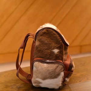 May include: A brown leather backpack with a unique spotted pattern. The backpack has a front pocket and two adjustable straps.