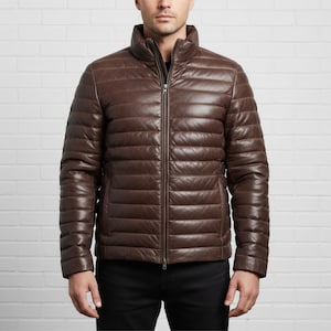 Puede incluir: Chaqueta de cuero marrón con diseño acolchado. La chaqueta tiene cuello alto y cremallera completa. Las mangas son largas y la chaqueta parece bien ajustada. El fondo es una pared de ladrillos blancos.