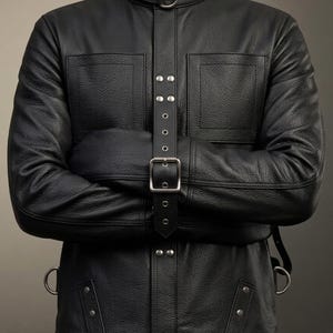 Può includere: Una giacca in pelle nera con colletto alto dotato di un anello a D in metallo, un cinturino verticale centrale con una grande fibbia argentata e numerosi rivetti metallici color argento. Il capo presenta due tasche sul petto e anelli laterali.