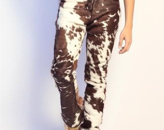 Op de afbeelding: Koeienhuid broek met een bruin en wit gevlekt patroon. De broek heeft een taille met trekkoord en wordt gedragen met beige laarzen. Het materiaal is zacht en harig.