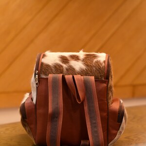 May include: A brown leather backpack with a cowhide flap and brown straps. The backpack has a zipper closure and is sitting on a wooden surface.