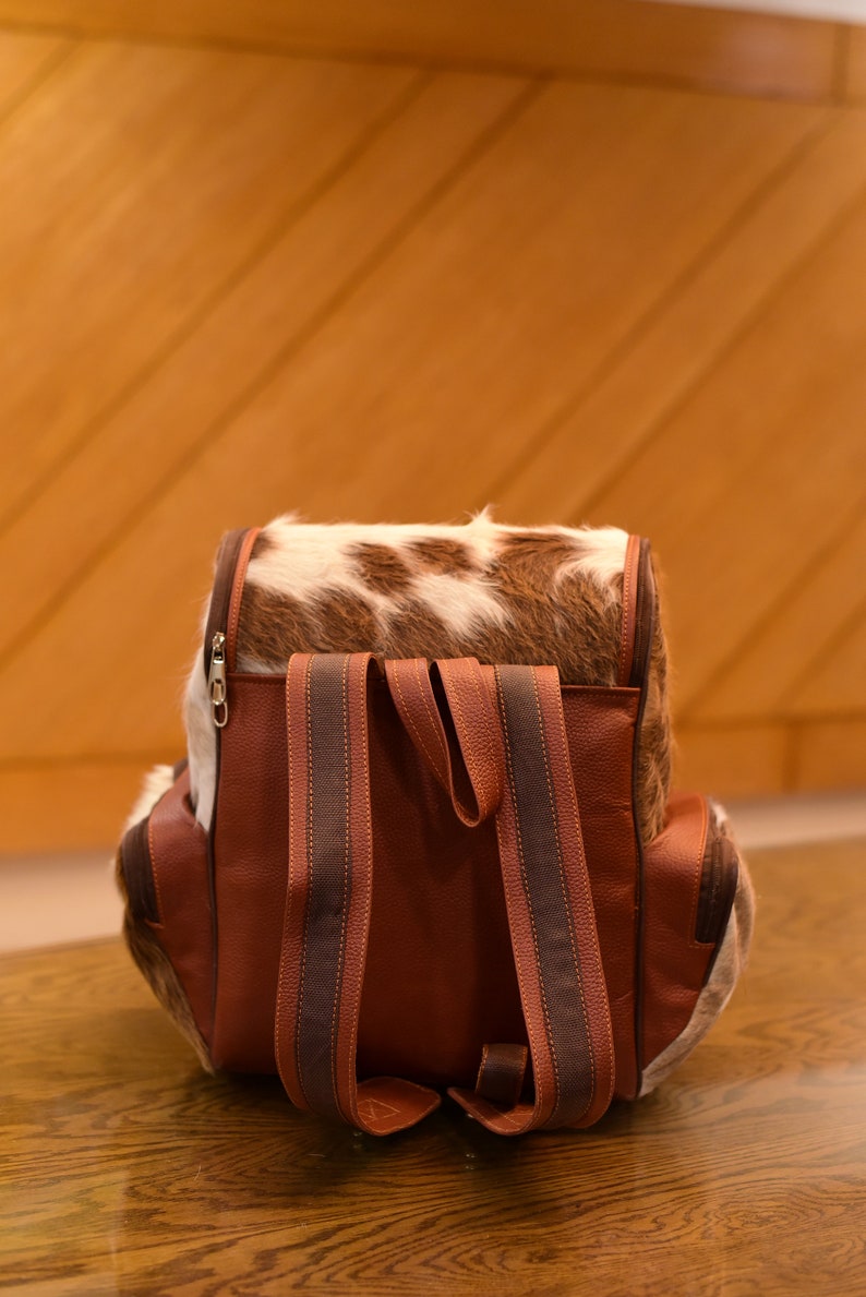 May include: A brown leather backpack with a cowhide flap and brown straps. The backpack has a zipper closure and is sitting on a wooden surface.