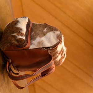May include: A brown leather backpack with a cowhide pattern. The backpack has a zipper closure and adjustable straps.