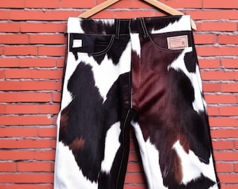 May include: Cowhide shorts with a black, brown, and white pattern, hanging on a wooden hanger. The shorts have a classic five-pocket design and a leather patch. The shorts have a white trim at the bottom.