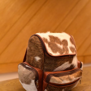May include: A brown and white cowhide backpack with brown leather accents and multiple zippered pockets.