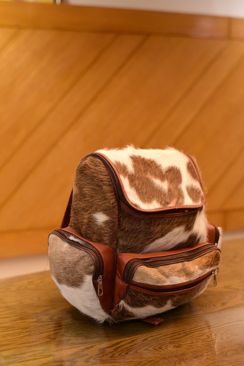 May include: A brown and white cowhide backpack with brown leather accents and multiple zippered pockets.