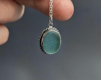 May include: A pendant necklace featuring an oval-shaped, frosted seafoam green piece of sea glass encased in a twisted silver metal frame. The pendant hangs from a delicate silver chain, showcasing a smooth, matte texture on the glass surface.