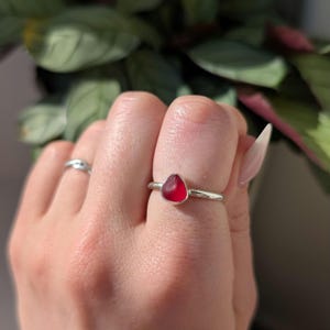 May include: A silver ring with a teardrop-shaped, deep red gemstone. The ring has a thin band and a bezel setting. The gemstone is smooth and polished. The ring is a simple, elegant piece of jewellery.