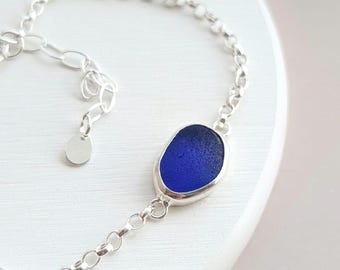 May include: A silver chain bracelet featuring a single, smooth, oval-shaped piece of deep cobalt blue sea glass set in a simple silver bezel. The bracelet includes a delicate link chain and a small, round, blank silver charm at the clasp.