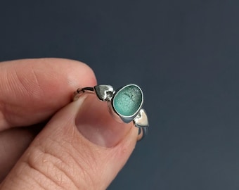 May include: A silver ring featuring a smooth, oval-shaped piece of sea glass in a soft teal-green color. The metal band is accented with two small, heart-shaped silver elements positioned on either side of the central sea glass setting.