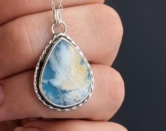 May include: A teardrop-shaped pendant necklace featuring a polished stone with swirling patterns of blue, white, and pale yellow. The stone is encased in a twisted silver metal frame and hangs from a delicate silver chain link necklace.
