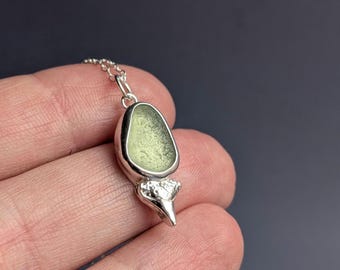May include: A pendant necklace featuring a smooth, pale green sea glass piece set in a polished silver bezel. The setting includes a textured, organic-shaped silver accent at the base, suspended from a delicate silver chain link.