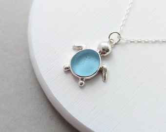 May include: A silver necklace featuring a turtle-shaped pendant with a smooth, light blue sea glass center. The pendant is attached to a delicate silver chain, showcasing a polished metal head, four flippers, and a circular shell setting.