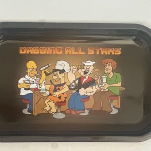 May include: Black rectangular rolling tray with rounded corners. The tray features a cartoon illustration of Homer Simpson, Fred Flintstone, Popeye, and Shaggy, with the text "Dabbing All Stars" in orange.