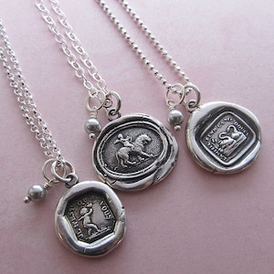 May include: Three silver necklaces with different charms. The charms are silver and have a wax seal design. One charm features a woman riding a lion, another features a swan, and the third features a child holding a flower.