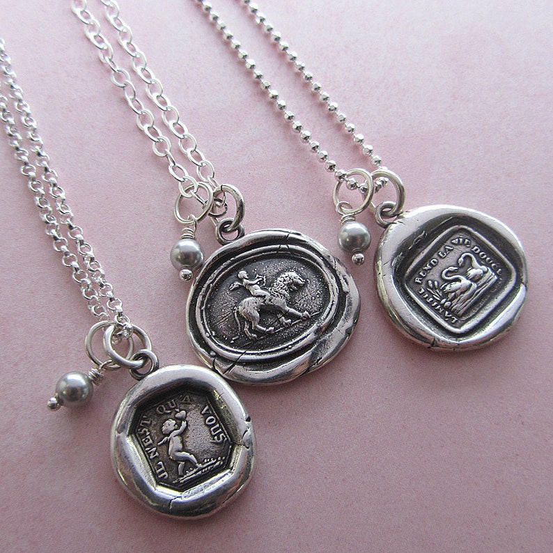 May include: Three silver necklaces with different charms. The charms are silver and have a wax seal design. One charm features a woman riding a lion, another features a swan, and the third features a child holding a flower.