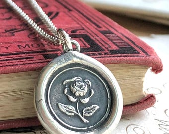 May include: A silver pendant necklace featuring a circular wax seal charm with a raised rose design. The charm has a dark, oxidized background and a polished silver border, suspended from a delicate silver chain link necklace.