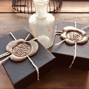 May include: Two black gift boxes with white string and a wax seal. The wax seals are brown and have a design of a letter and leaves. The boxes are sitting on a wooden surface.