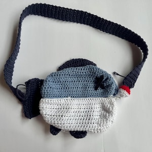 May include: A handmade crochet fish-shaped crossbody bag. The bag features a blue and white body with a dark blue strap and accents. A small red and white bobber charm hangs from the strap. The fish has a black 'X' on its eye.