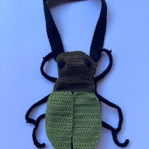 May include: A handmade crochet bag in the shape of a beetle. The bag features a dark green head and light green body, with black antennae and a strap. The bag is designed to be worn over the shoulder.