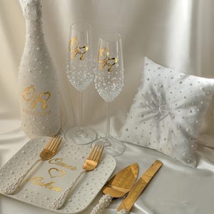 Könnte beinhalten: Eine Sammlung von Hochzeitsaccessoires, darunter eine dekorierte Champagnerflasche, zwei Sektgläser, ein Ringkissen, ein Torten-Servierset und ein Teller mit Gabeln. Die Artikel sind weiß mit Perlenakzenten und goldfarbenen Schriftzügen. Auf der Champagnerflasche steht das Datum 09.05.2025.