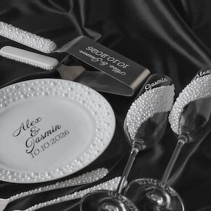 Könnte beinhalten: Elegantes Hochzeitsset mit einem weißen Teller, Tortenheber, Gabeln und Sektgläsern, alle mit Kunstperlen verziert. Teller und Heber sind mit Namen und Datum graviert. Das Set ist auf dunklem, strukturiertem Untergrund angeordnet.