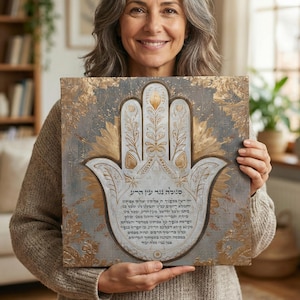 May include: A square, grey and gold Hamsa hand art piece. The Hamsa hand is white with gold accents and intricate designs. The background is a distressed grey with gold floral patterns. Text is visible on the artwork.