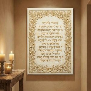 May include: A rectangular wall art piece with gold-colored text and a decorative olive branch border. The text is in Hebrew. The artwork is displayed on a textured, off-white background, and is mounted on a wall. Candles are on a wooden table in the foreground.