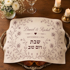 May include: A rectangular cream-colored fabric challah cover featuring a dark brown floral vine border. The center displays the text Daniel & Rachel, a heart icon, the date SEPTEMBER 5, 2026, and the Hebrew text Shabbat Shalom in dark brown ink.