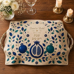 Könnte beinhalten: Eine rechteckige, cremefarbene Stoffabdeckung für ein Tablett mit verzierten goldfarbenen Metallgriffen, verziert mit einem blau-türkisfarbenen Granatapfel- und Rankenmuster. Der Text lautet Daniel & Rachel, MAY 12, 2024, sowie hebräisch Shabbat V'Yom Tov.