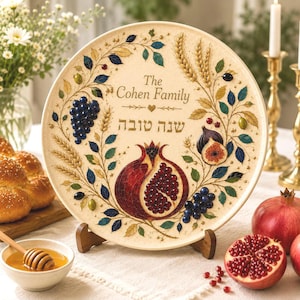 May include: A round decorative ceramic plate featuring a central pomegranate illustration, clusters of blue grapes, figs, and wheat stalks. The text reads The Cohen Family and in Hebrew letters. The plate has a cream background with gold accents.
