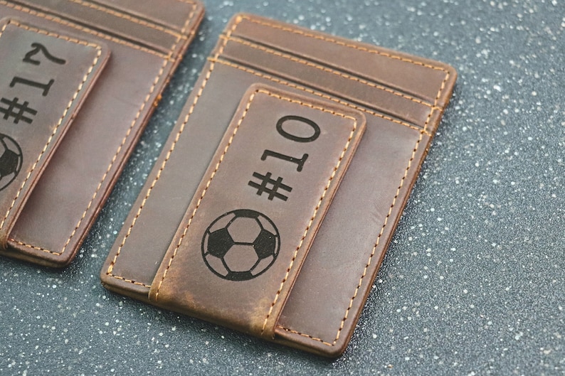 Puede incluir: Dos carteras de cuero marr&oacute;n con un clip para dinero y un dise&ntilde;o de bal&oacute;n de f&uacute;tbol. Las carteras est&aacute;n grabadas con los n&uacute;meros "#17" y "#10".