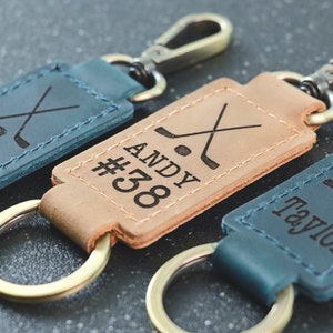 May include: Leather keychains in teal and tan with hockey stick and puck graphics. Each keychain has a name or number: "#55", "Andy #38", and "Taylor". They have gold-colored hardware and a key ring.